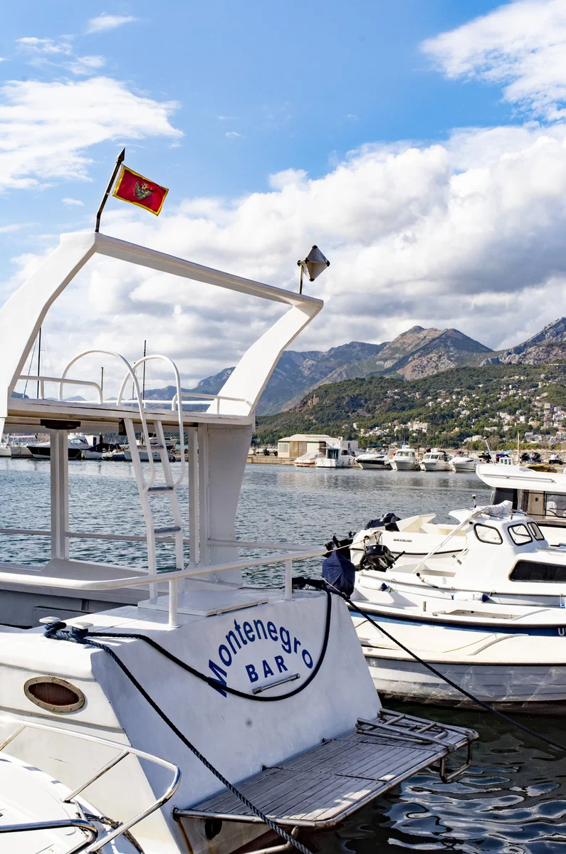 White boat with Montenegro Bar written on it and a Montenegrin flag at Bar marina, with mountains and other boats in the background
