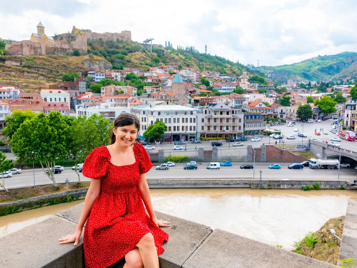 Woman in a red dress sitting by the Mtkvari River with a panoramic view of Tbilisi’s old town, Narikala Fortress, and historic churches in the background. Photo taken from Metekhi chutch.